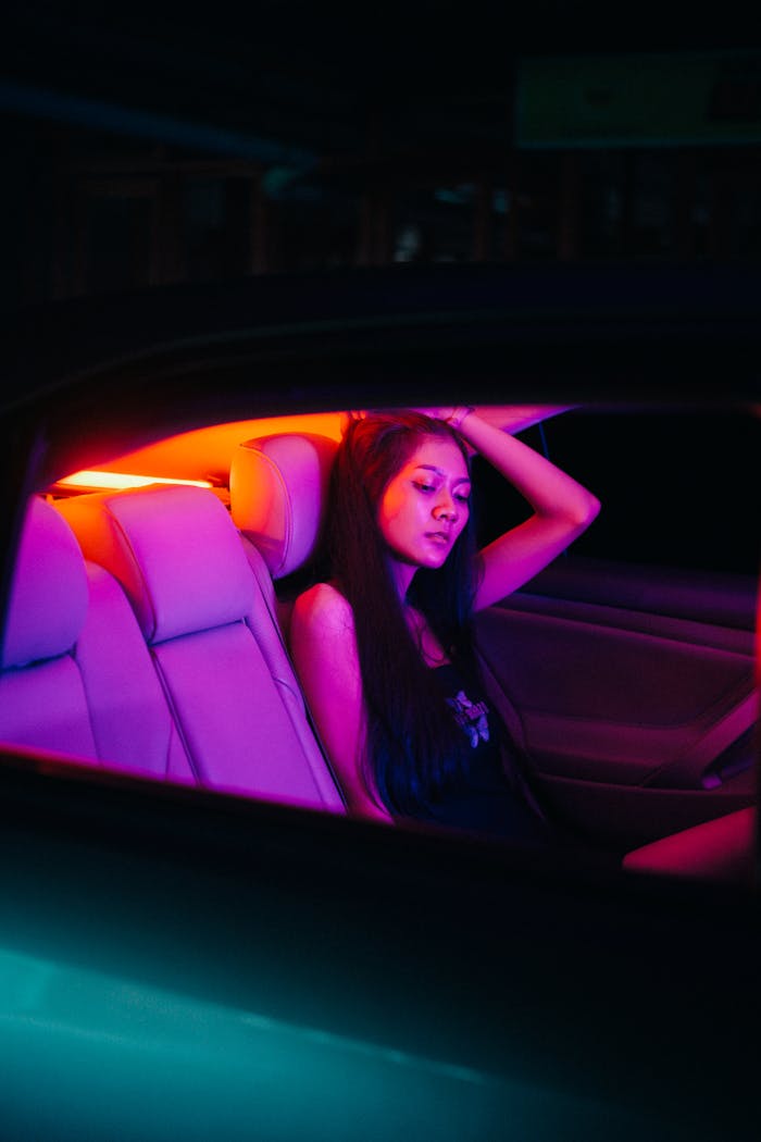 An Asian woman relaxes in a car interior illuminated by vibrant neon lights at night.